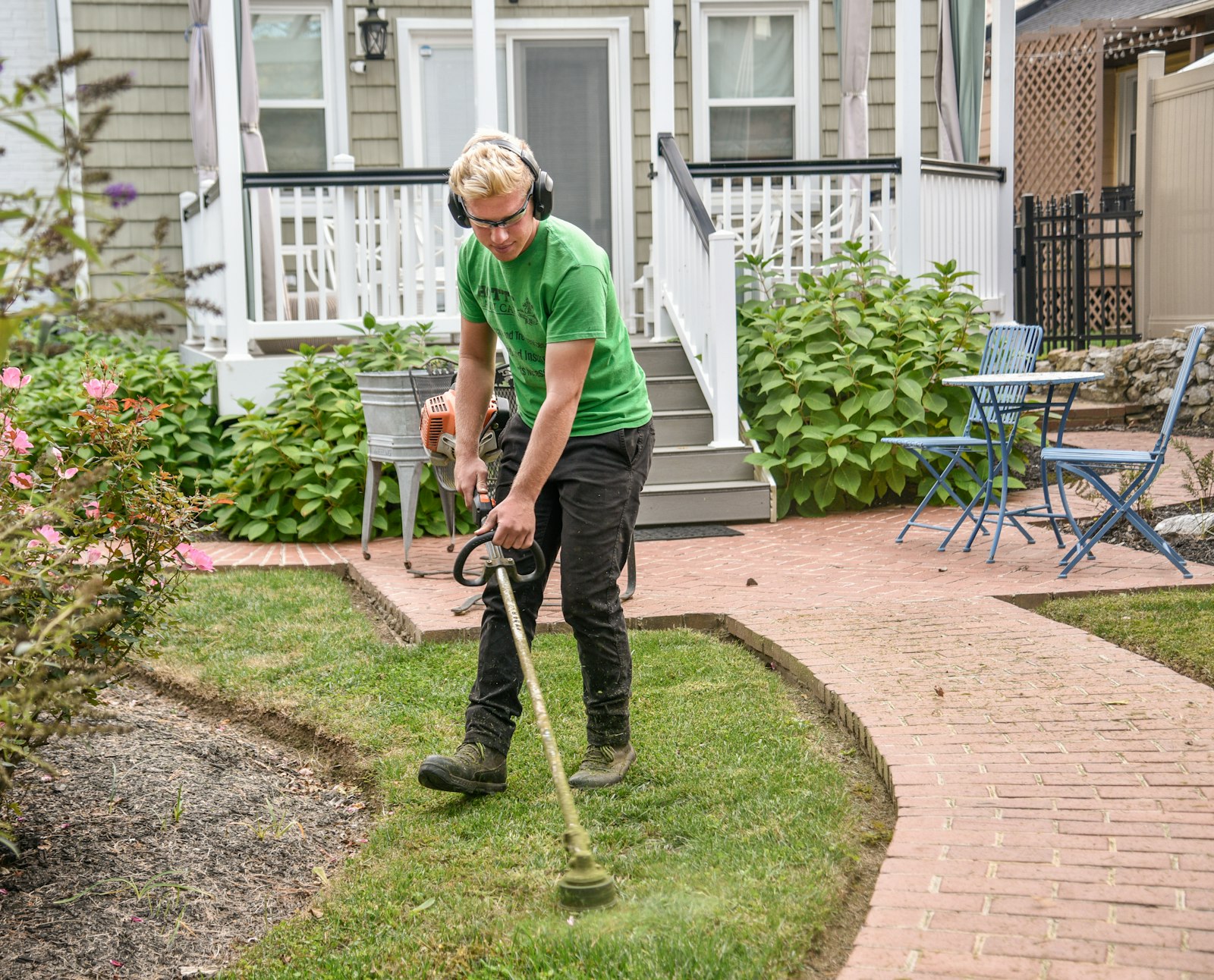A landscaper edge-trimming a lush green lawn beside a brick paver walkway